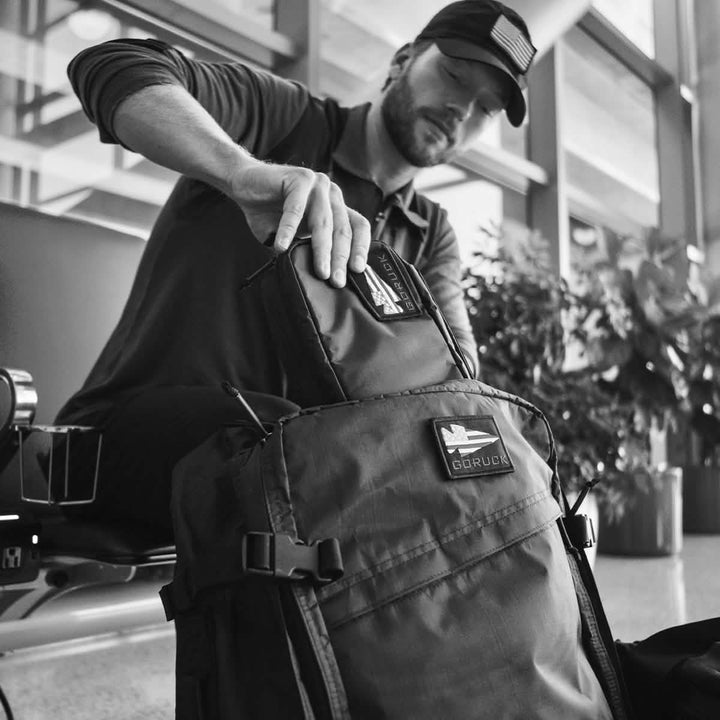 A bearded man in a cap meticulously packs his black GORUCK bag with the GR1 Field Pocket - Dyneema, a smaller pouch that fits snugly inside. Seated and focused, he organizes his belongings against the backdrop of a modern building interior accented by large windows and greenery. Black and white photo.