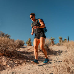 Under a clear blue sky, a person in athletic gear and sunglasses hikes a rugged trail, enjoying the comfort of GORUCK's Merino Challenge Socks - Ankle. Carrying a backpack, they navigate the path surrounded by dry shrubs and desert plants.