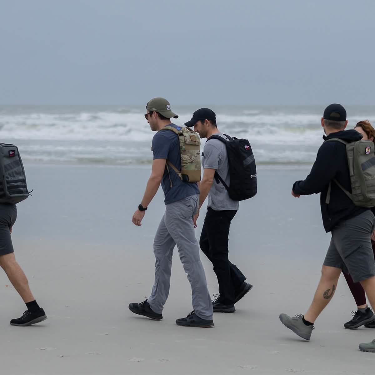 On a cloudy day, five people stroll along a sandy beach near the ocean, each wearing backpacks and dressed in casual outdoor clothing. Every step they take is comfortably cushioned by their GORUCK Merino Challenge Socks - Ankle, ideal for seaside adventures.