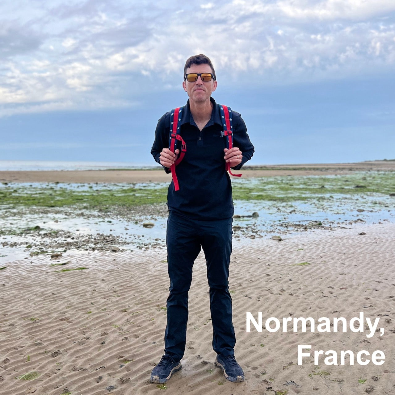 Man wearing Men's Rucking Shirt - Merino Wool in black with a red backpack stands on a sandy beach under cloudy skies. Text: 