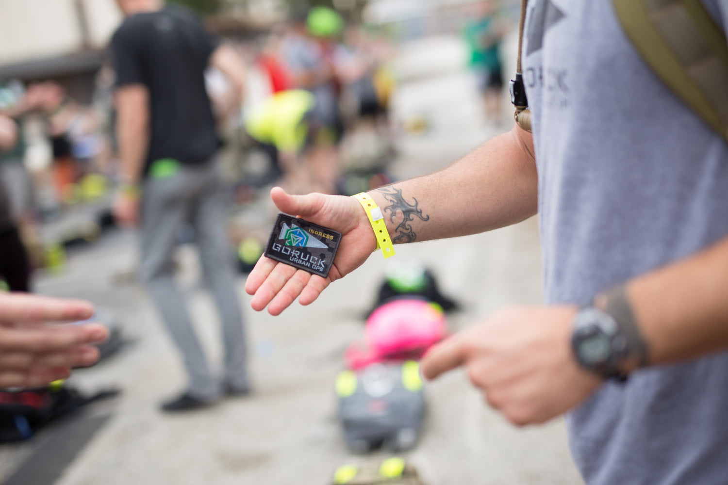 A person holding a patch at an outdoor event, with people and gear blurred in the background.