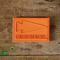Orange GORUCK patch with arrowhead logo on worn wooden workbench surface