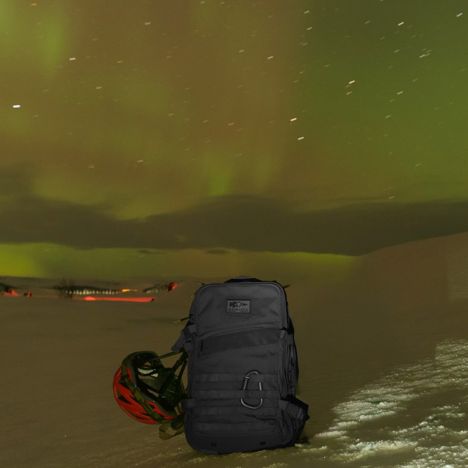 The Rucker - Long Range rucksack and a helmet sit on snowy ground beneath green northern lights, ready for long-distance rucking adventures under the night sky.