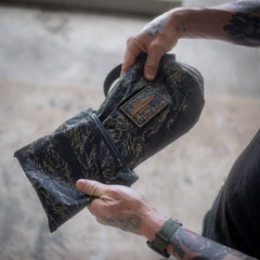 Tattooed hands holding the Packable Bullet Ruck – Ripstop ROBIC®, a black patterned pouch with a patch that says “ESTABLISH.” Featuring AquaGuard zippers, this lightweight day bag is expertly designed for both style and practicality.
