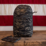 A camouflage-patterned Packable Bullet Ruck - Ripstop ROBIC® backpack and pouch sit on a wooden surface with a red and white flag behind, featuring AquaGuard zippers for extra protection—making it the perfect lightweight day bag for any adventure.