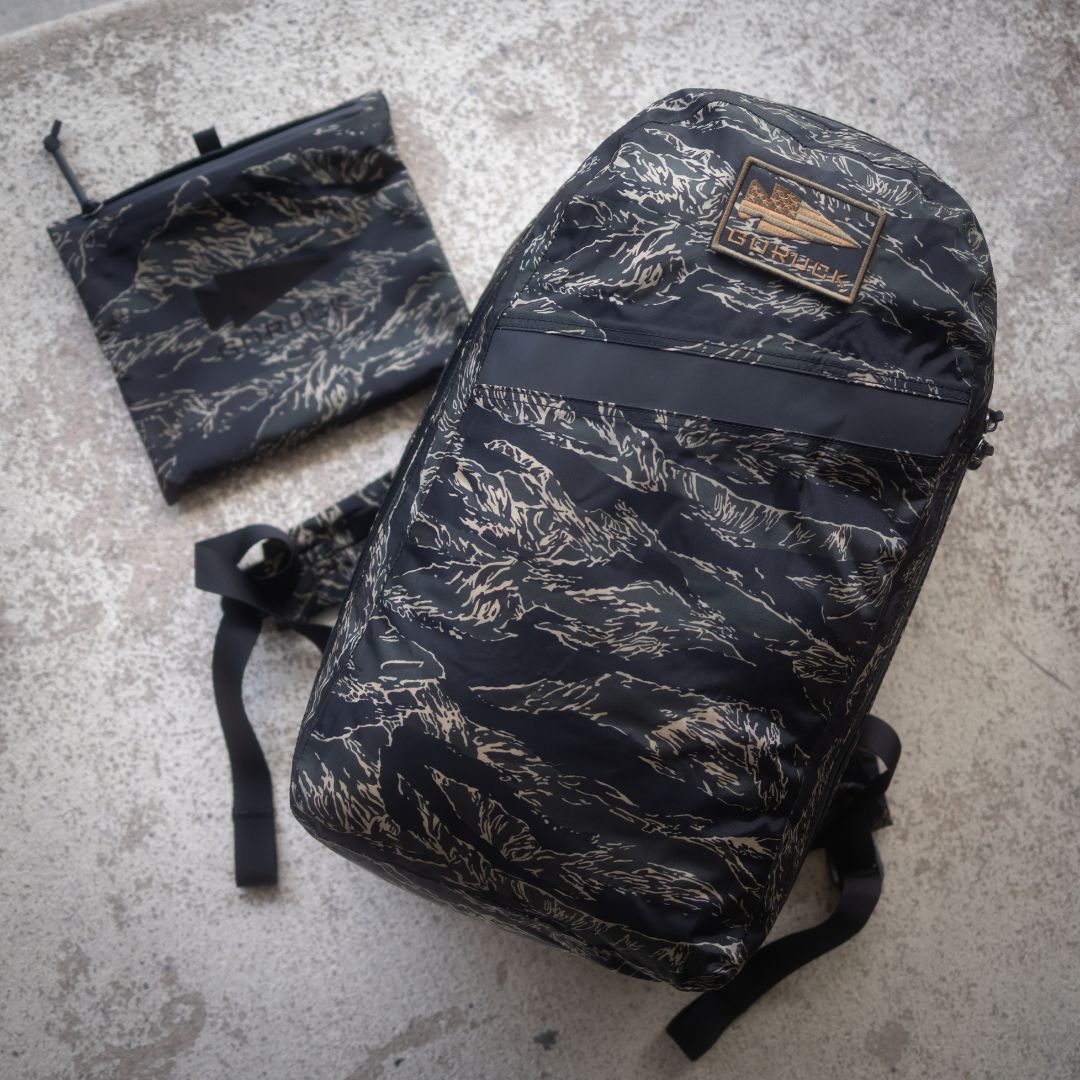 The Packable Bullet Ruck - Ripstop ROBIC® in black camo pattern with AquaGuard zippers and matching pouch sits atop a textured concrete surface—offering the perfect lightweight day bag set for any adventure.