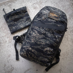 The Packable Bullet Ruck - Ripstop ROBIC® in black camo pattern with AquaGuard zippers and matching pouch sits atop a textured concrete surface—offering the perfect lightweight day bag set for any adventure.