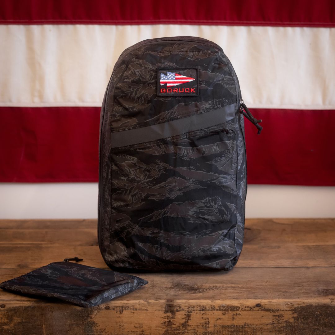 A black camo Packable Bullet Ruck - Ripstop ROBIC® with a US flag patch sits on a wooden table, an American flag in the background. Featuring AquaGuard zippers, this lightweight day bag is both durable and stylish—perfect for everyday adventures.