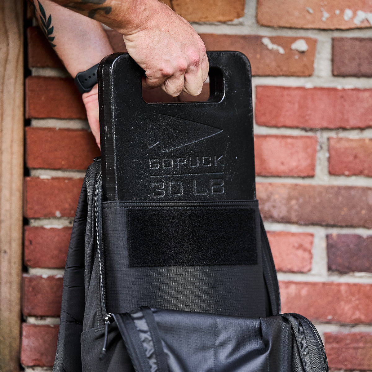A person puts a 30 lb GORUCK weight plate into a black Basic Rucker® backpack in front of a brick wall.