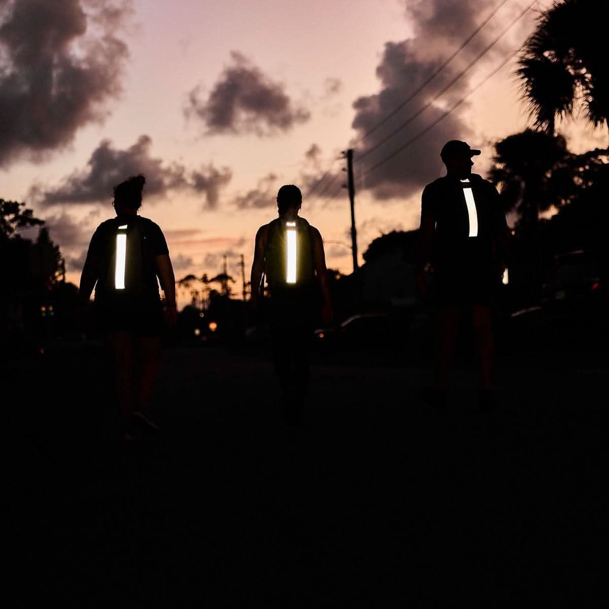 Three people wearing reflective gear walk outdoors at dusk, rucking together as the sun sets behind palm trees in the background. Each carries a Basic Rucker backpack fitted with a ruck plate, blending fitness and adventure into their evening stroll.