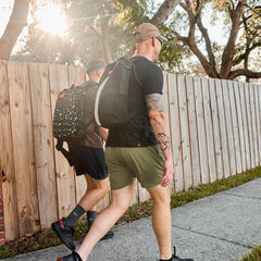 Two men with backpacks, including a Basic Rucker®, walk on a sidewalk next to a wooden fence on a sunny day.