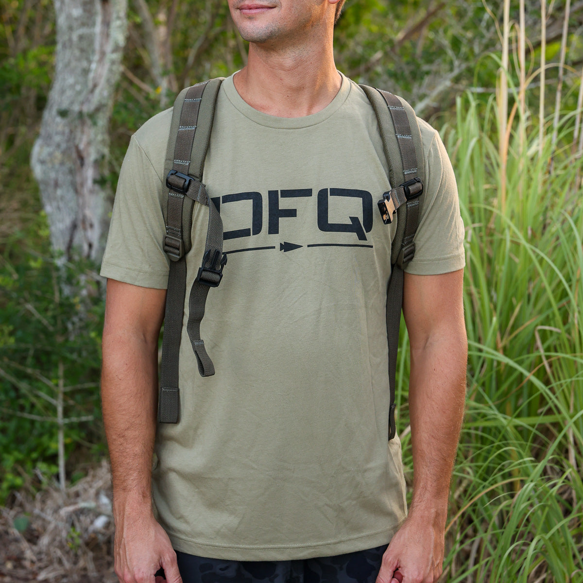A man wearing a khaki DFQ Tee - Tri-Blend and a backpack stands outdoors in front of green foliage.