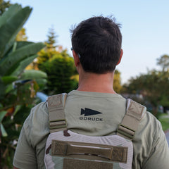 A man outdoors with greenery is seen from behind wearing a DFQ Tee - Tri-Blend motivational shirt and a camouflage backpack.