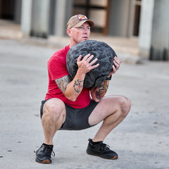 A man in a red shirt and cap squats outdoors with the Sand Medicine Ball, making it ideal for portable workouts or adding variety to your home gym routine.