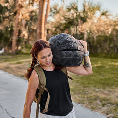 A woman carries a Sand Medicine Ball on her shoulder outdoors, ideal for portable training or home gym workouts, with trees and grass in the background.