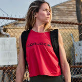 Woman outdoors wearing red GORUCK tank top and rucking backpack, chain-link fence behind
