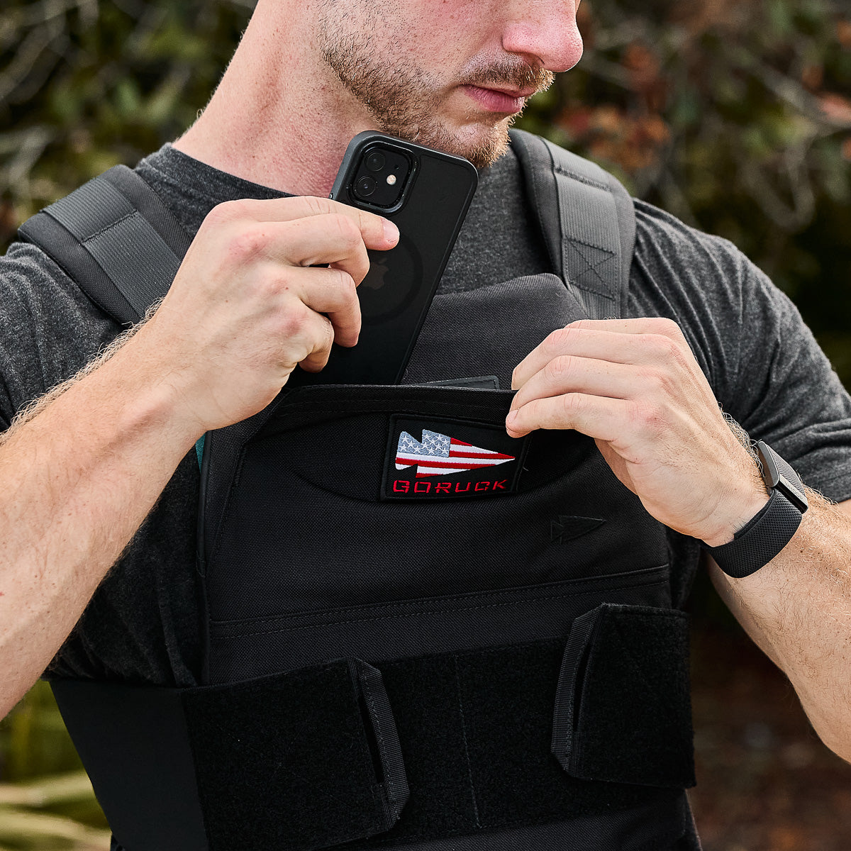 Man inserting a phone into a chest pocket on a black tactical vest with an American flag patch.