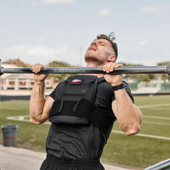 A man wearing the Rucking Weight Vest with Curved Ruck Plates does a pull-up on an outdoor bar, with a field visible in the background.