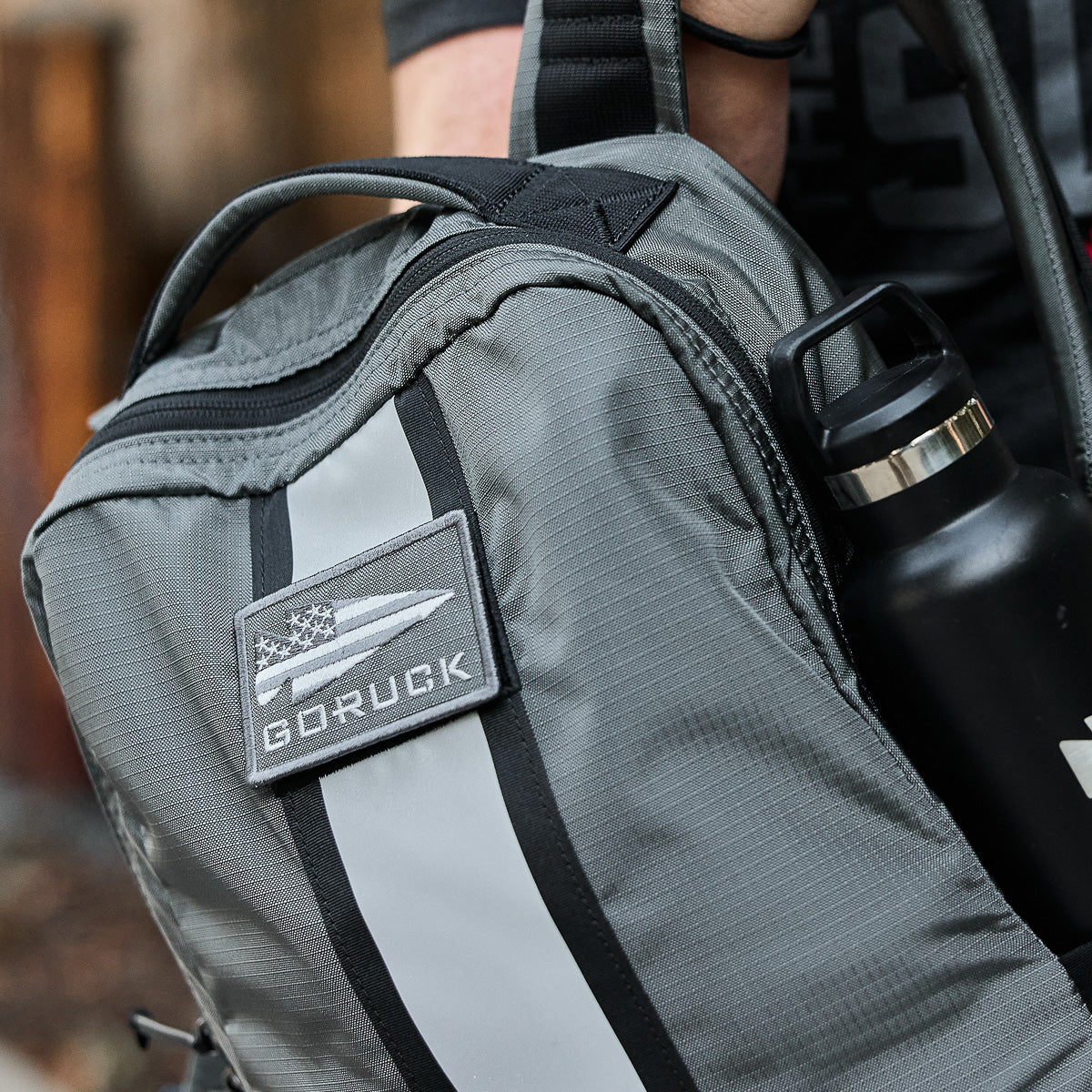 A close-up of a gray GORUCK backpack with a patch and a black water bottle in the side pocket.