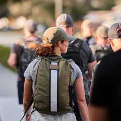 A group of people wear Basic Rucker® weighted rucksacks and caps as they walk outdoors on a sunny day.