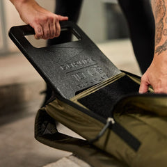 A person puts a 15 lb "Born Primitive" workout plate into an olive green Basic Rucker®, turning it into a weighted rucksack for training.