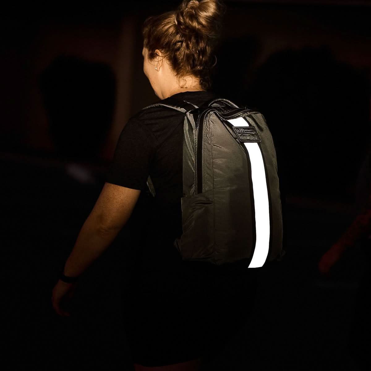 A person with a Basic Rucker backpack, which features a bright reflective strip, walking in a dimly lit area.