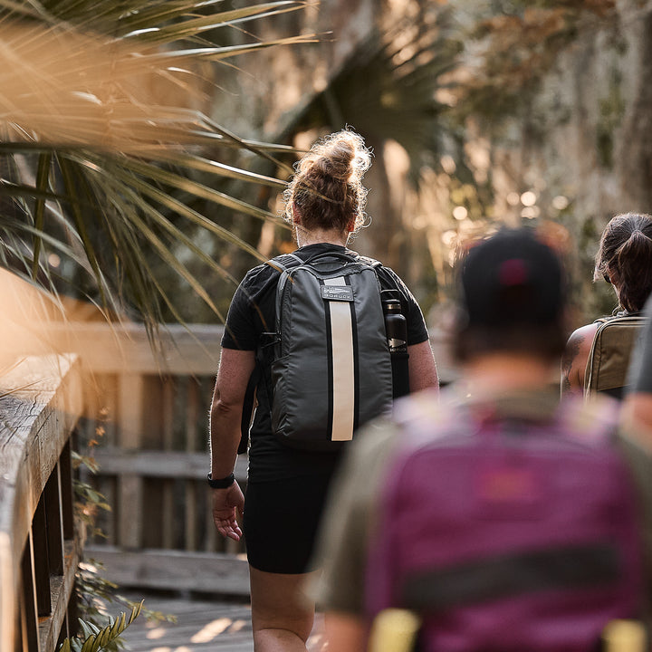 A person carries the Basic Rucker® on a wooden trail surrounded by trees, with others following.