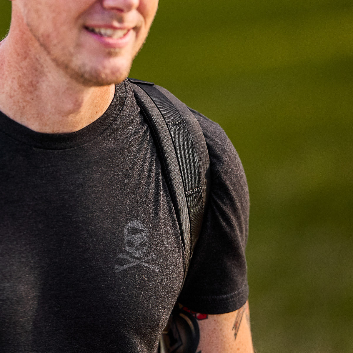 Man in a black t-shirt with a backpack strap, smiling, standing outdoors with a blurred green background.