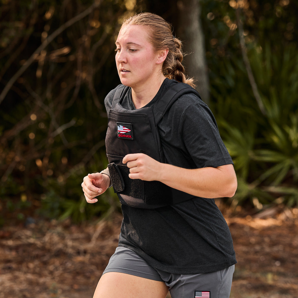 A woman runs outdoors in athletic gear and a Rucking Weight Vest with Curved Ruck Plates, surrounded by lush greenery—perfect for rucking enthusiasts.