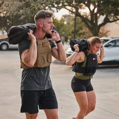 Two people wearing Rucking Weight Vests with Curved Ruck Plates carry sandbags on their shoulders while rucking outdoors.