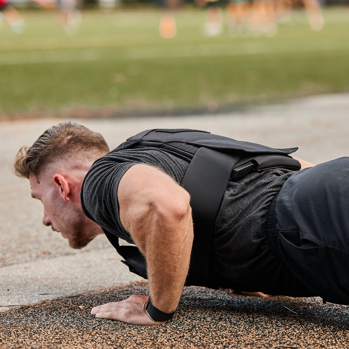 A man does push-ups outdoors in a Rucking Weight Vest loaded with Curved Ruck Plates, grass and people blurred in the background—ideal gear for rucking enthusiasts pushing their limits.
