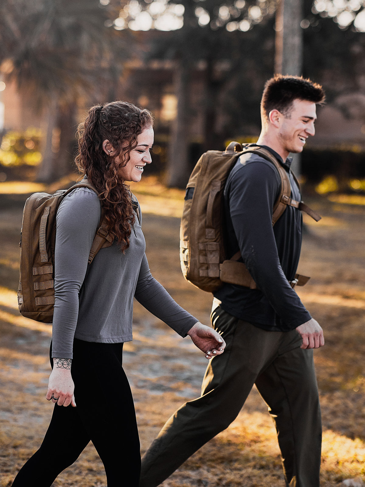 Two people walk outdoors wearing backpacks, smiling and dressed in casual athletic clothing.
