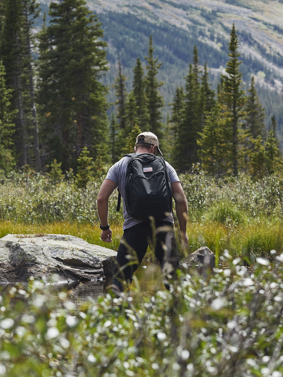 A man with a backpack stands near rocks in a forested mountain landscape, facing away.