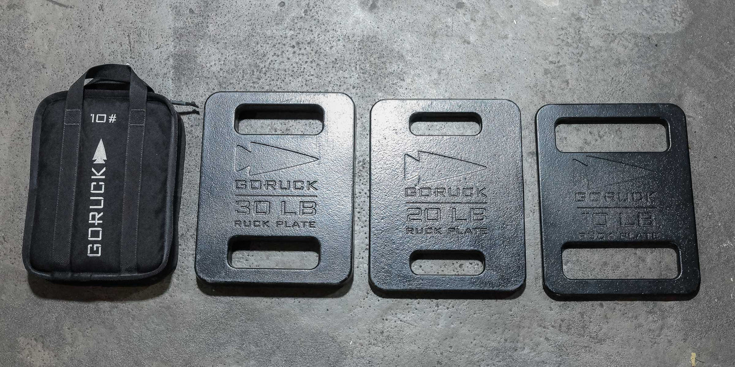 Four GORUCK ruck plates, labeled 10, 30, 20, and 10 pounds, on a concrete floor.