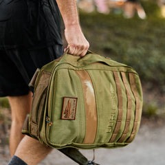 Someone outdoors carries the Rucker 4.0, an olive green rucking backpack, by its top handle while wearing black shorts.
