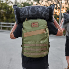 A person carries a camo sandbag and wears the Rucker 4.0 weighted training pack outdoors at sunset.