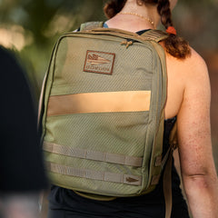 A person outdoors, seen from behind, wears an olive green Rucker 4.0 backpack with ruck plate compatibility as sunlight shines on the bag.