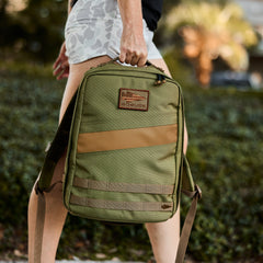 A person walks outdoors near greenery, holding the Rucker 4.0 in olive green by the top handle. This gym rucksack is ruck plate compatible, making it ideal for outdoor fitness adventures.
