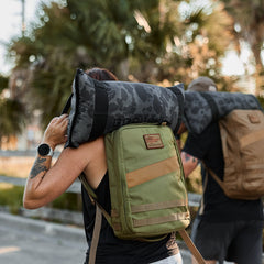 Two people carry Rucker 4.0 backpacks and camo-patterned sandbags on their shoulders outdoors by palm trees.