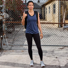 Woman wearing blue sleeveless top and black leggings with backpack standing in front of chain link fence