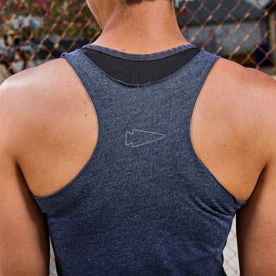 A person in a blue Women's Racerback Tank - Tri-Blend by GORUCK stands from behind, ready for a workout in front of a chain-link fence, with blurred foliage softly painting the background.