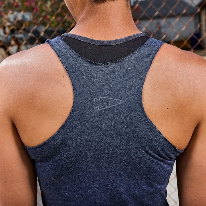 A person in a blue Women's Racerback Tank - Tri-Blend by GORUCK stands from behind, ready for a workout in front of a chain-link fence, with blurred foliage softly painting the background.