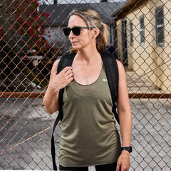 woman wearing olive green tank top, black backpack, and sunglasses standing by chain-link fence outdoors