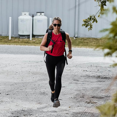 Wearing a Women’s Performance Tank - ToughMesh from GORUCK, black leggings, and sunglasses, a person jogs briskly along a gravel path with a backpack. With greenery and propane tanks in the background, they move with precision as if training for a trek through the Italian Alps.