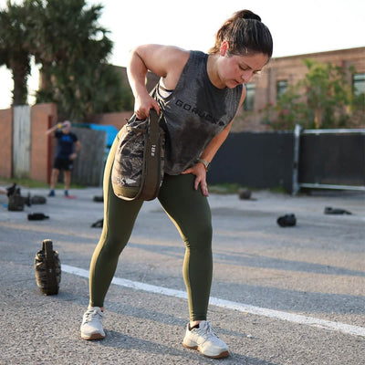 A woman in workout attire bends over while holding a weighted bag outdoors on a sunny day. She is wearing a gray Women’s Performance Tank made with GORUCK's ToughMesh™ fabric, green leggings, and white sneakers. Other workout equipment and participants can be seen in the background, creating a scene reminiscent of the Italian Alps.