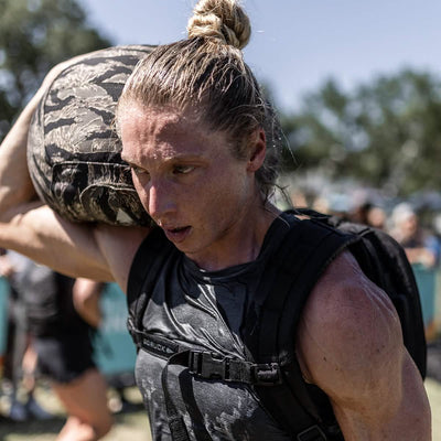 A determined athlete with a topknot hoists a hefty sandbag onto their shoulder during an outdoor competition. Wearing the Women's Performance Tank by GORUCK, crafted with ToughMesh™ fabric, they push through the event, as blurred spectators and trees surround them, evoking the rugged landscapes of the Italian Alps.