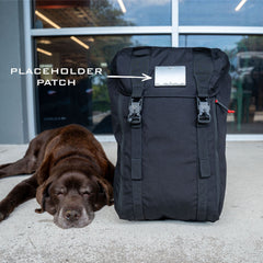 A black M26 PREORDER // Q4 2026 ETA field pack with a "placeholder patch" label sits next to a sleeping brown dog on a concrete floor, featuring a bombproof laptop compartment for added durability.