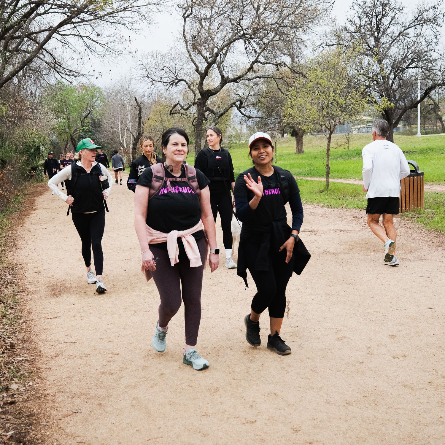 Two women in stylish GORUCK MOTHERUCKER Women's Tee - Tri-BlendX smile and wave at the camera while walking on a tree-lined park trail.