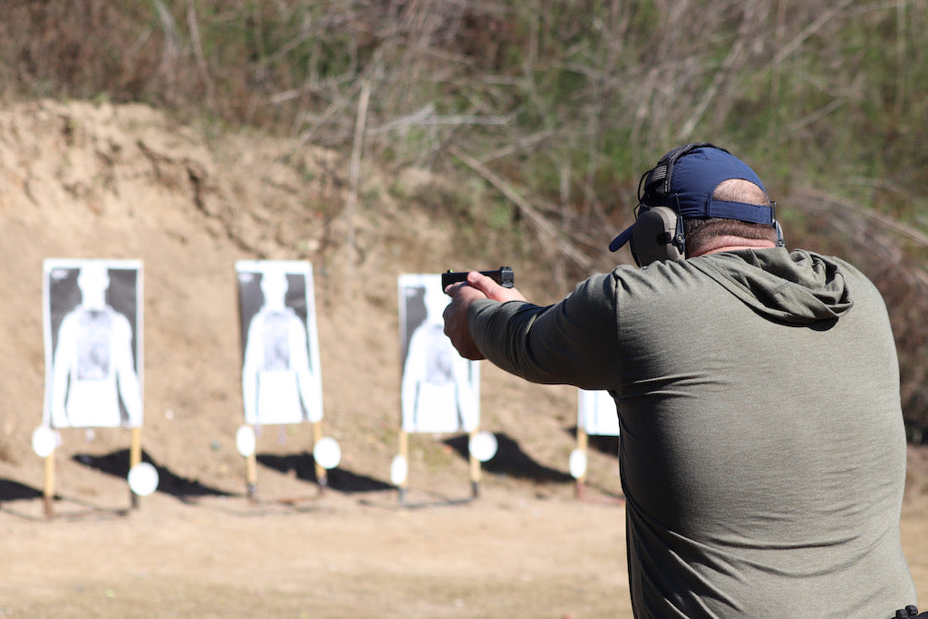 Man aiming a handgun at paper targets on an outdoor shooting range, wearing ear protection and a cap.