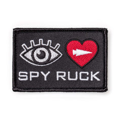 The Patch - Eye Heart Spy Ruck is a rectangular, eye-catching patch with an eye, red heart with white arrow, and "SPY RUCK" text—perfect for espionage style fans.